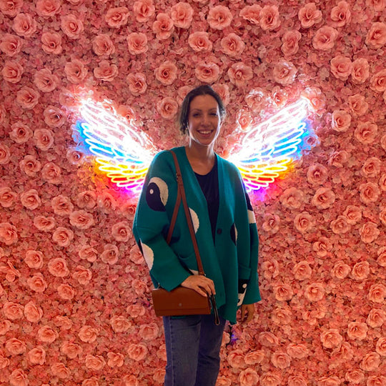 Woman standing in front of a floral wall with neon butterfly wings holding an archive crossbody bag
