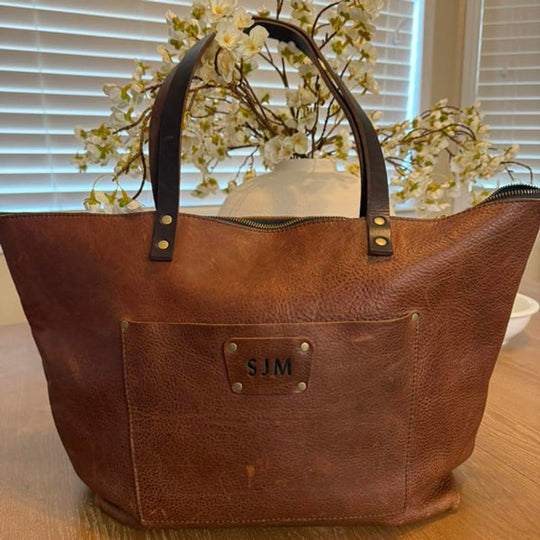 Leather tote bag with 'SJM' monogram on a wooden surface with flowers in the background.