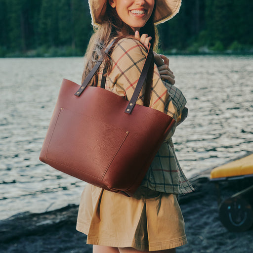 A woman outdoors by the water wearing a wide-brim hat and carrying a large brown leather tote bag over her shoulder. featured image