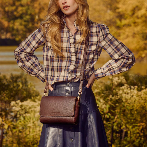 A model outdoors wearing a leather crossbody bag with a matching strap, styled with a beige and navy plaid shirt and a dark skirt. featured image