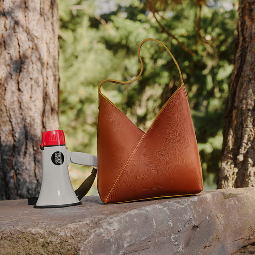 A brown pebbled leather shoulder bag with a single strap, styled on a stone ledge outdoors next to a white and red megaphone, with trees and green foliage in the background. featured image
