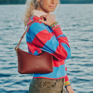 A woman wearing a colorful red and blue striped sweater stands by the water, carrying a brown pebbled leather shoulder bag with a small gold logo.