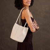 Bone Classic | Model wearing sleeveless dark brown dress carrying petite bucket shaped tote bag with matching leather handles against dark grey background
