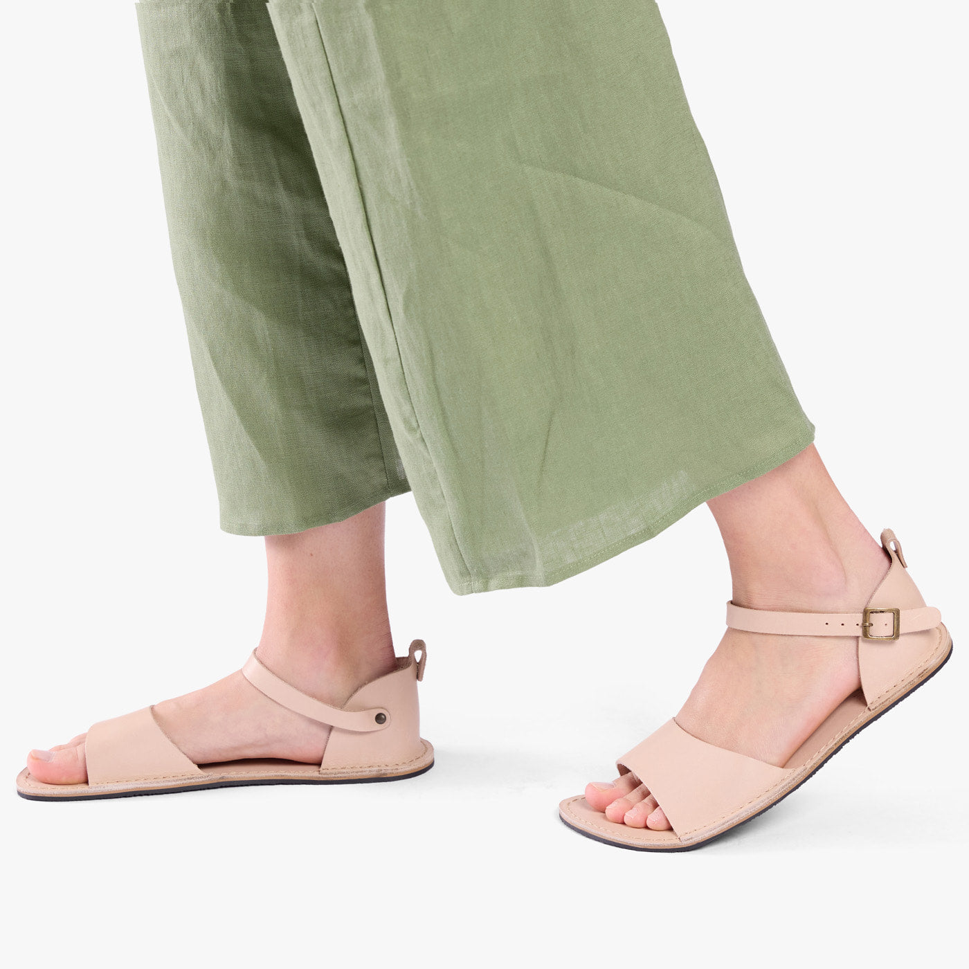 Crema | Close-up of a person wearing light green cropped pants and cream flat sandals with an ankle strap and buckle, shown against a plain background.