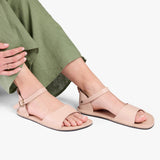 Crema | Close-up of a person wearing light green cropped pants and cream flat sandals with an ankle strap and buckle, shown against a plain background.