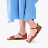 Miele | Person wearing blue dress and tan flat sandals with an ankle strap and buckle, shown against a plain background.