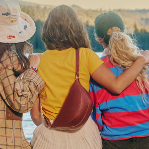 Three women sitting together outdoors by a lake, viewed from behind. The central woman wears a brown leather crescent-shaped sling bag across her yellow shirt.