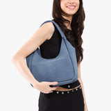 Denim Medium | A model holding a u-shaped, flat-bottomed leather shoulder bag with two outside pockets