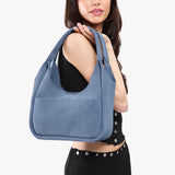Denim Medium | A model holding a u-shaped, flat-bottomed leather shoulder bag with two outside pockets