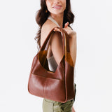 Nutmeg Medium | A model holding a u-shaped, flat-bottomed leather shoulder bag with two outside pockets
