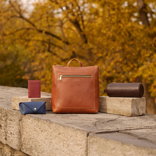 A leather backpack with a top handle and front zip pocket sits on a stone ledge outdoors, surrounded by matching leather accessories including a pouch, a card holder, and a cylindrical case. featured image