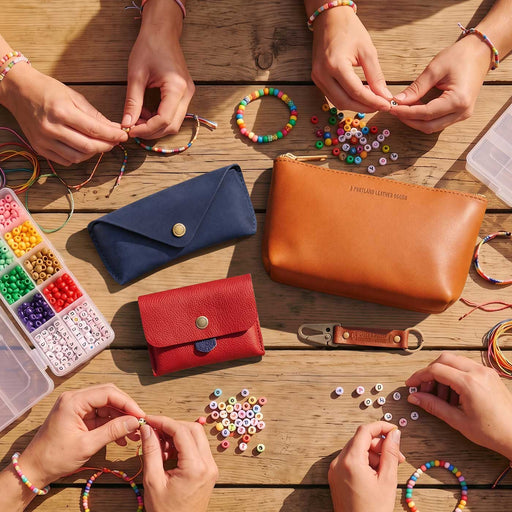 A top-down view of several hands crafting beaded bracelets and necklaces around a wooden table, with jewelry-making supplies including colorful beads, thread, a blue and red leather wallet, a tan leather pouch, and a bead organizer. featured image