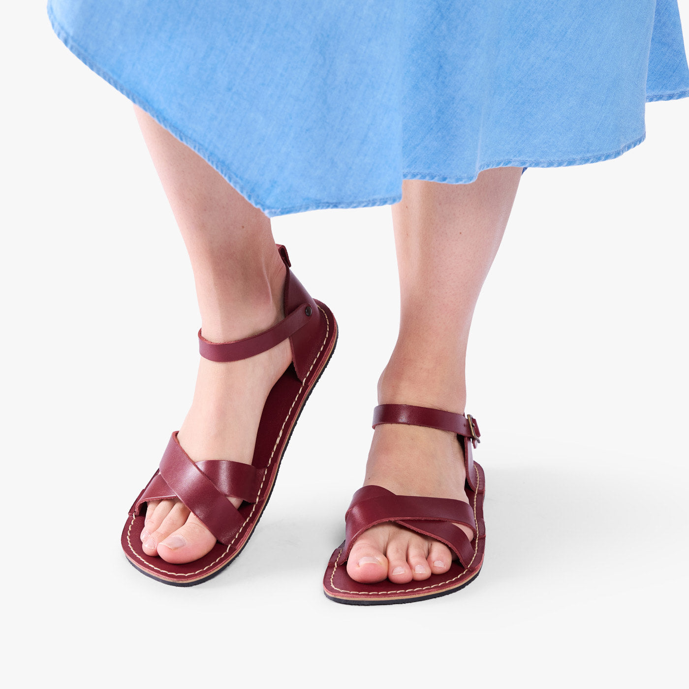 Marsala | Close-up of model wearing light blue dress and reddish brown flat sandals with an ankle strap and buckle, shown against a plain background
