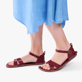 Marsala | Close-up of model wearing light blue dress and reddish brown flat sandals with an ankle strap and buckle, shown against a plain background