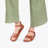 Miele | Close-up of model wearing light green pants and brown flat sandals with an ankle strap and buckle, shown against a plain background