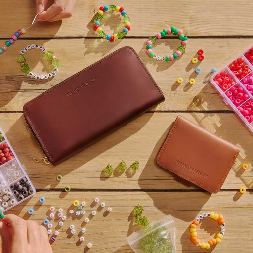 Two embossed leather zip-around wallets, shown in a styled flat-lay on a wooden table surrounded by colorful beaded bracelets, beads, and small accessories. One wallet is dark brown and the other is tan. featured image
